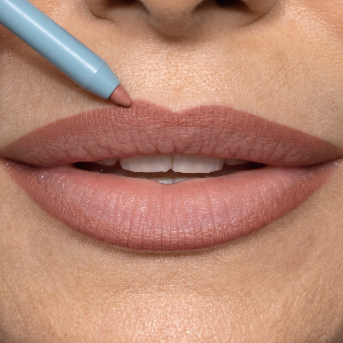 A close-up of a person’s lips as they apply nude lip liner to define the lip shape. The lip liner pencil is touching the upper lip, and the lips are slightly parted, showing the teeth.