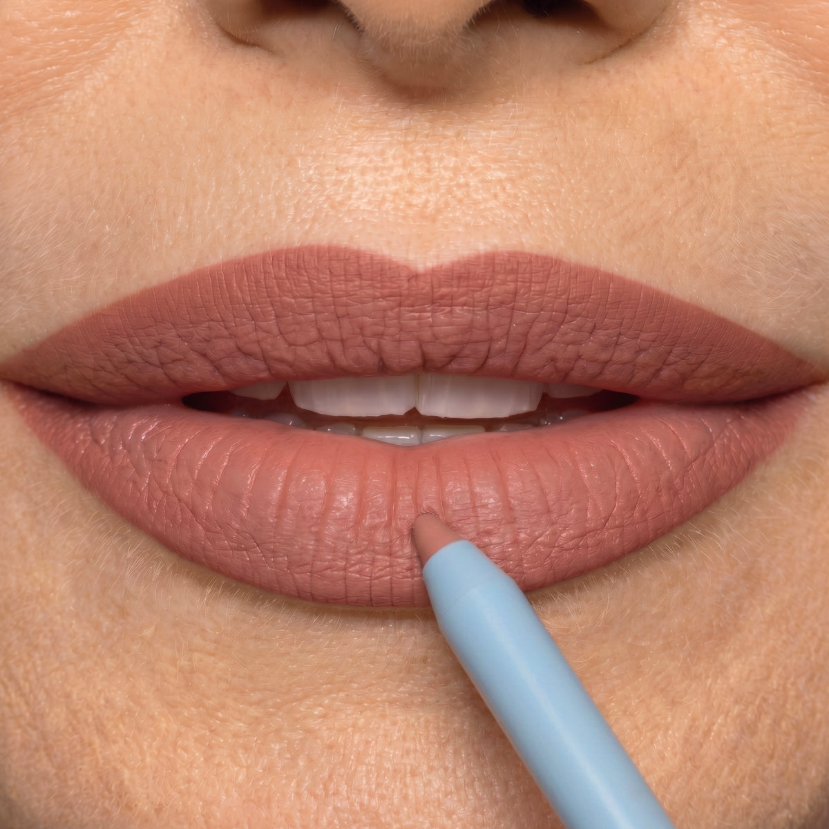 A close-up of a person applying natural nude lip liner to their lips, which are slightly parted, revealing their teeth. The lip liner pencil is held near the lower lip.