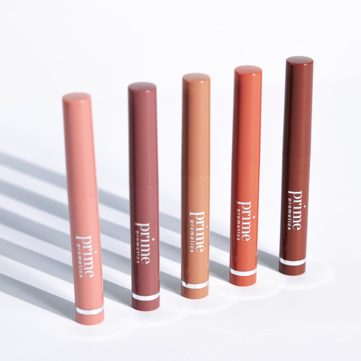 Five Prime Cosmetics lip crayon tubes in various shades of pink, mauve, nude, orange, and brown are standing upright in a row on a white surface with soft shadows.