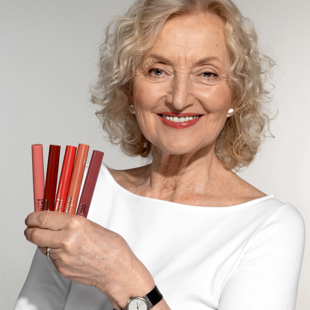 Smiling older woman with curly blonde hair in a white top holds five different shades of lipstick tubes, fanned out in her hand, against a neutral background.