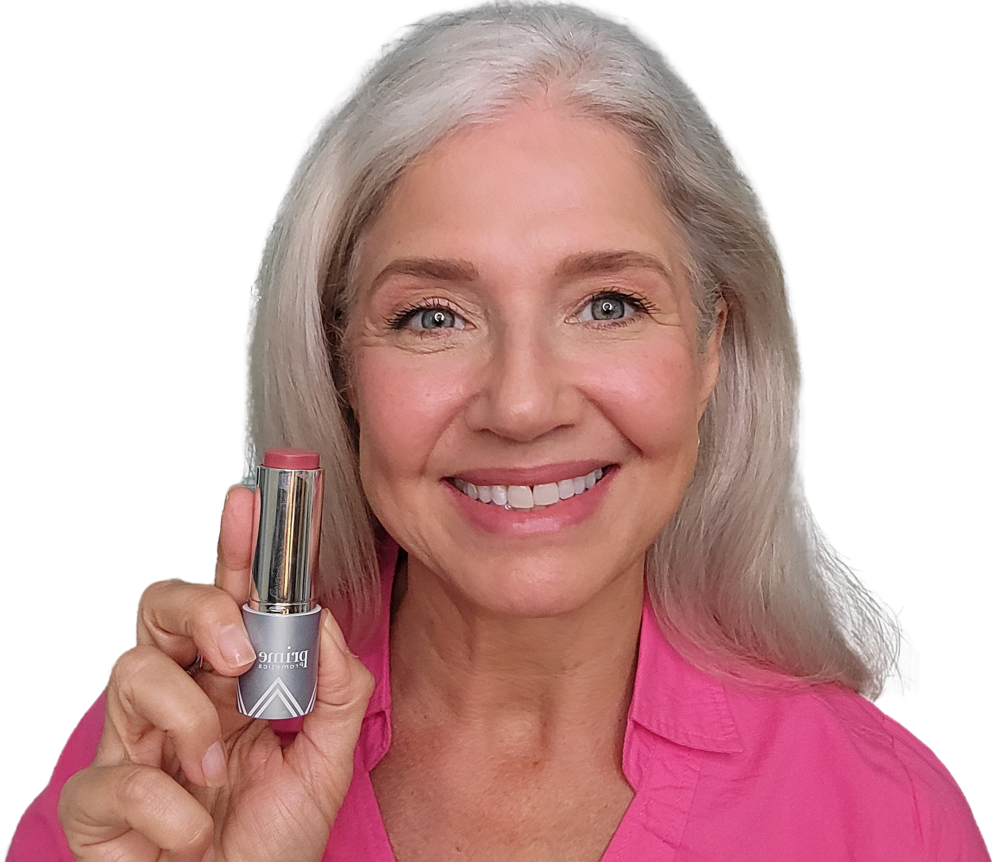 Smiling older woman with long gray hair, wearing a pink shirt, holds up a tube of pink lipstick in her right hand against a plain white background.