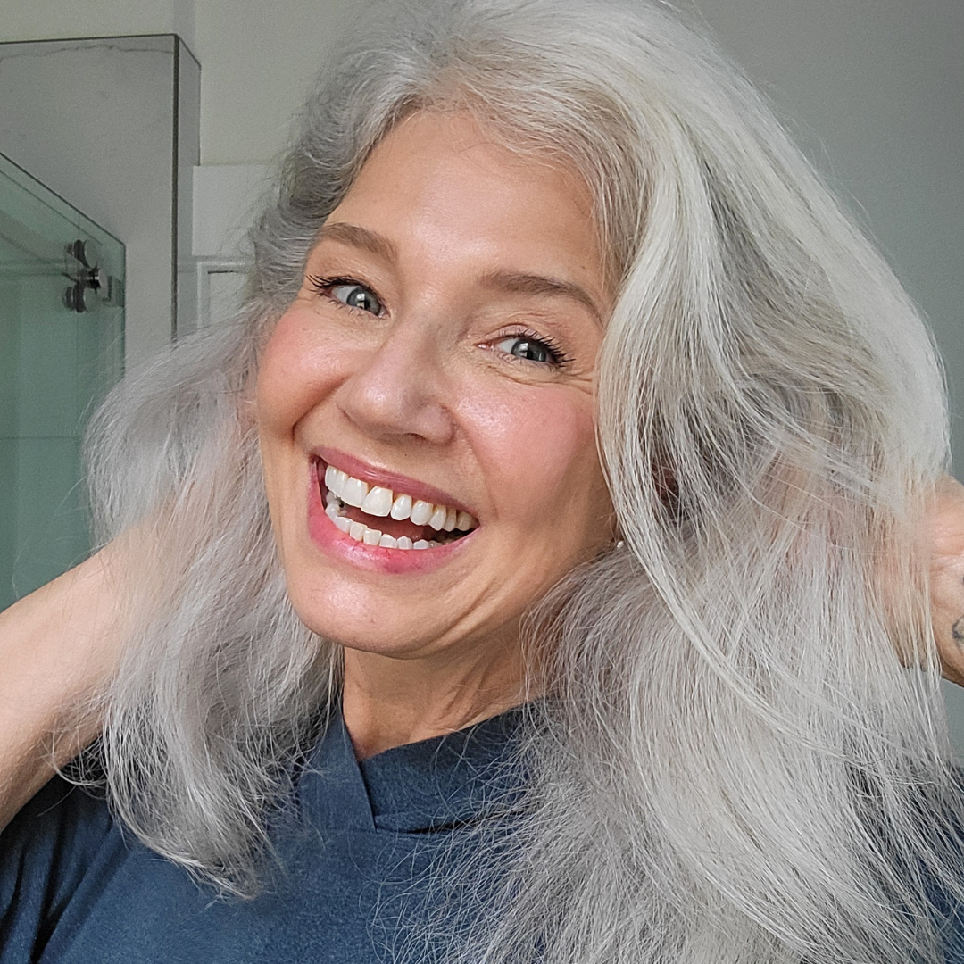 A smiling older woman with long, wavy gray hair holds her hair back with both hands, wearing a dark top and standing in a bright bathroom.