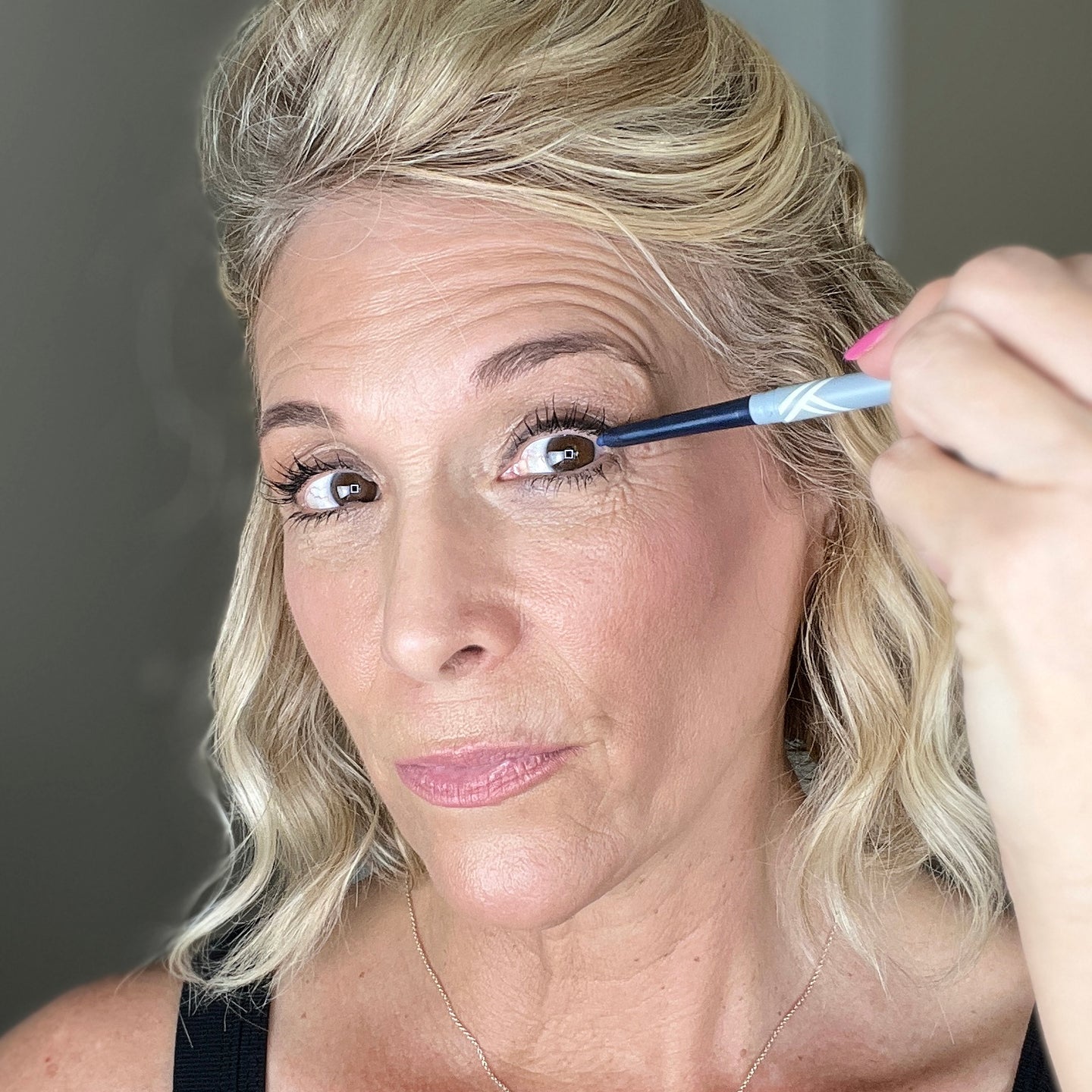 A woman with blonde hair applies eyeliner to her upper eyelid while looking into the camera. She has wavy hair and is wearing a black top. The background is neutral and slightly out of focus.