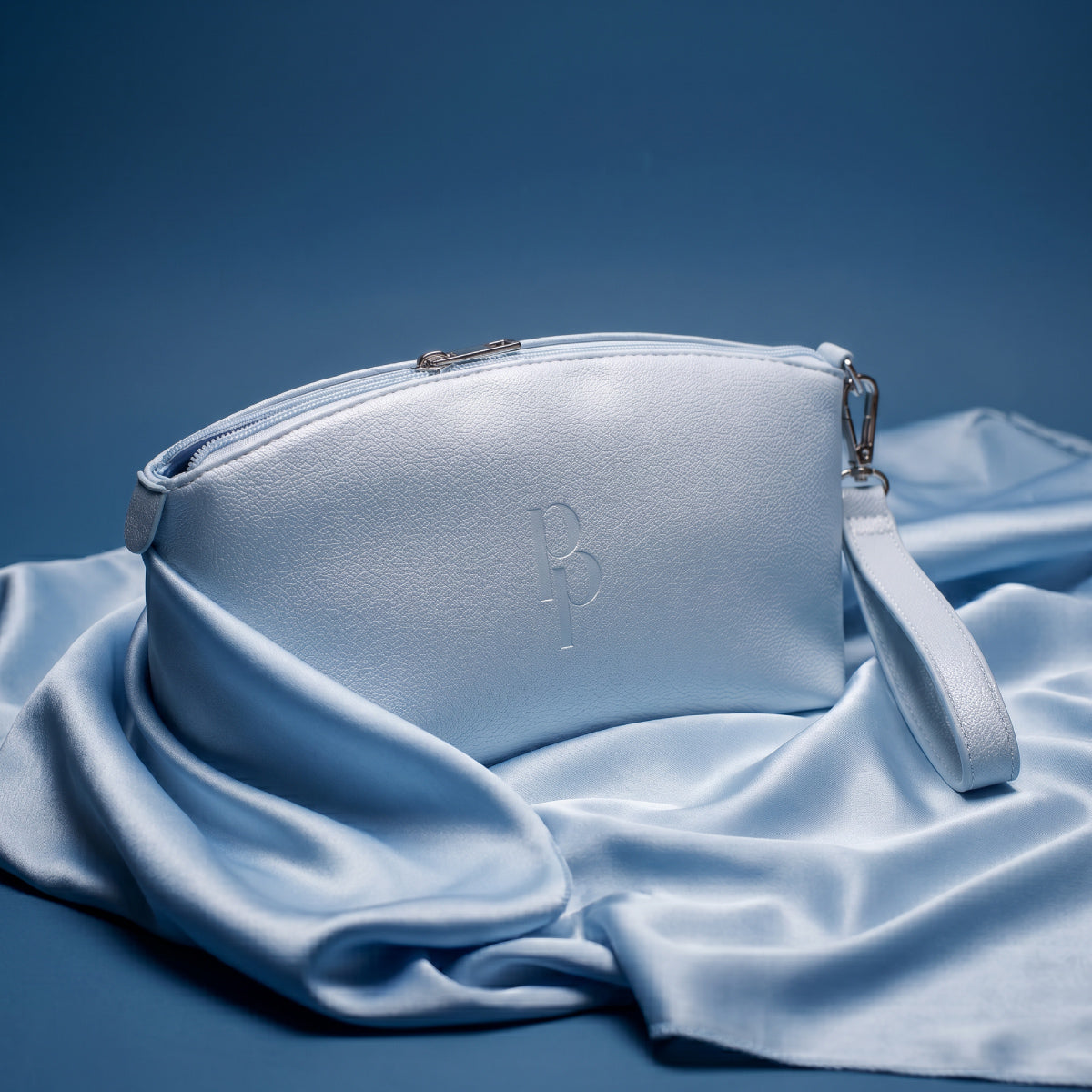 A light blue Makeup Bag by PrimePrometics™, crafted from elegant PU leather and featuring a convenient wrist strap, rests on matching blue satin fabric against a coordinating blue background—making it the ultimate beauty bag for any occasion.