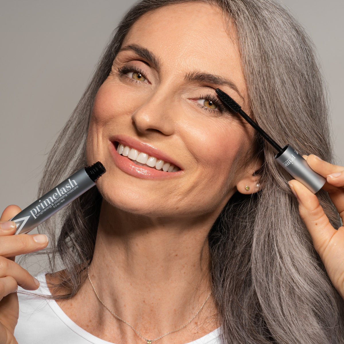A smiling woman with long gray hair applies mascara to her eyelashes, holding a silver tube labeled primelash in one hand and a mascara wand in the other. She is wearing a white top and delicate jewelry.