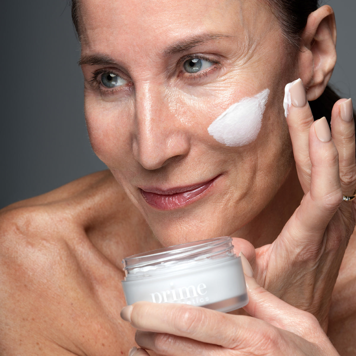 A middle-aged woman with clear skin applies a white cream to her cheek, holding an open jar labeled prime in her other hand. She smiles gently and appears to be in a skincare routine.