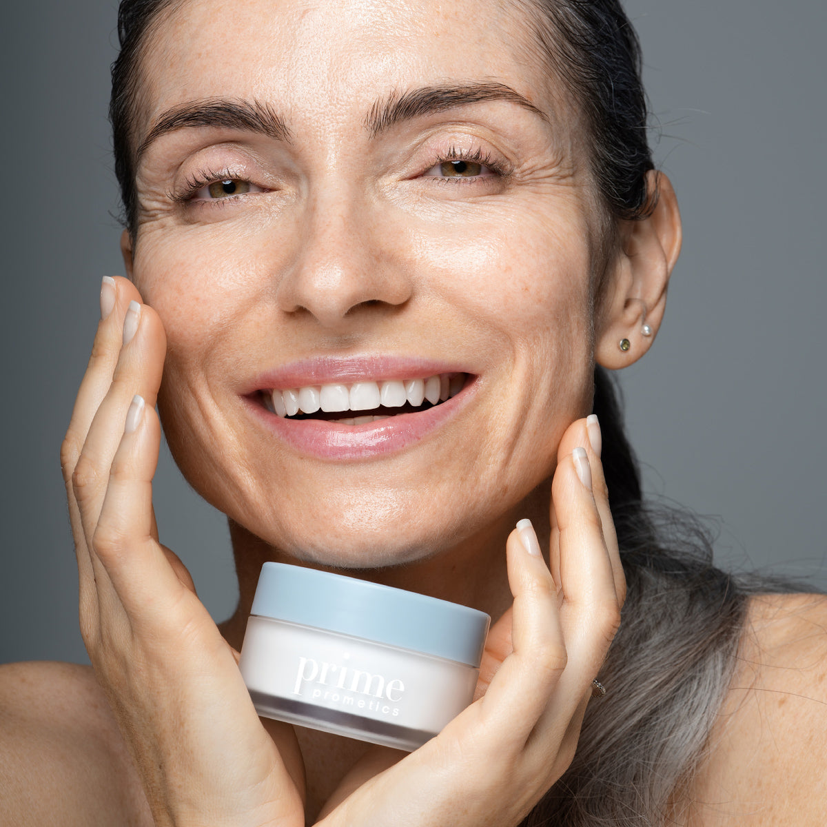 A smiling woman with glowing skin holds a jar of Prime Cosmetics cream near her face, gently touching her cheek with her other hand. She appears happy and confident, with minimal makeup.
