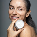 Mature woman smiling and holding an open jar of Prime Prometics Miracle Cream, showing its rich whipped texture