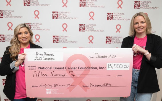Two women stand smiling, holding a large pink check for $15,000 made out to the National Breast Cancer Foundation. The background features repeated foundation logos with pink ribbons.
