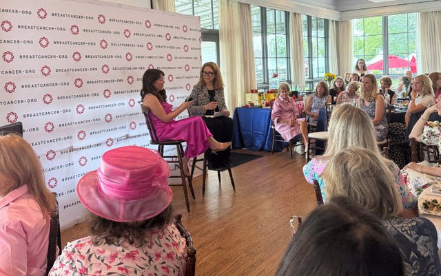 Two women sit on high chairs on a stage, speaking in front of an audience at a BreastCancer.org event. One woman wears a pink dress, and the audience is dressed in spring attire, with many women wearing hats.