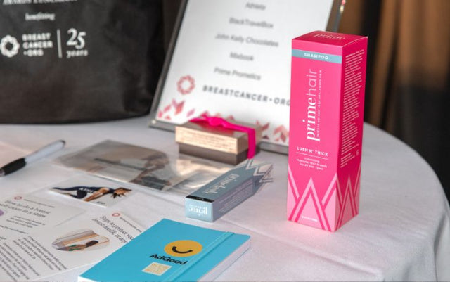 A table displays various products, including a pink Primehair shampoo box, business cards, brochures, a blue AdGood booklet, and a framed sign with a breast cancer awareness logo in the background.