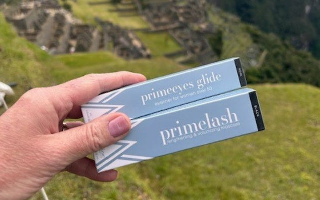 A hand holds two cosmetics boxes labeled primeyes glide and primelash with ancient stone ruins and green hills visible in the blurred background.