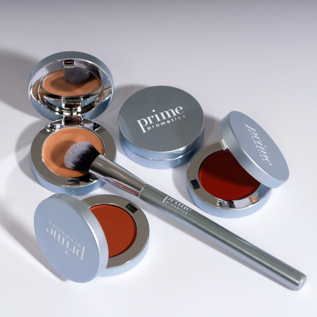 Drei offene, runde Make-up-Dosen in silbernen Hüllen, eine davon mit einem Pinsel darin, sowie zwei geschlossene Dosen. Außerdem liegt ein Make-up-Pinsel mit silbernem Griff schräg über den Dosen.