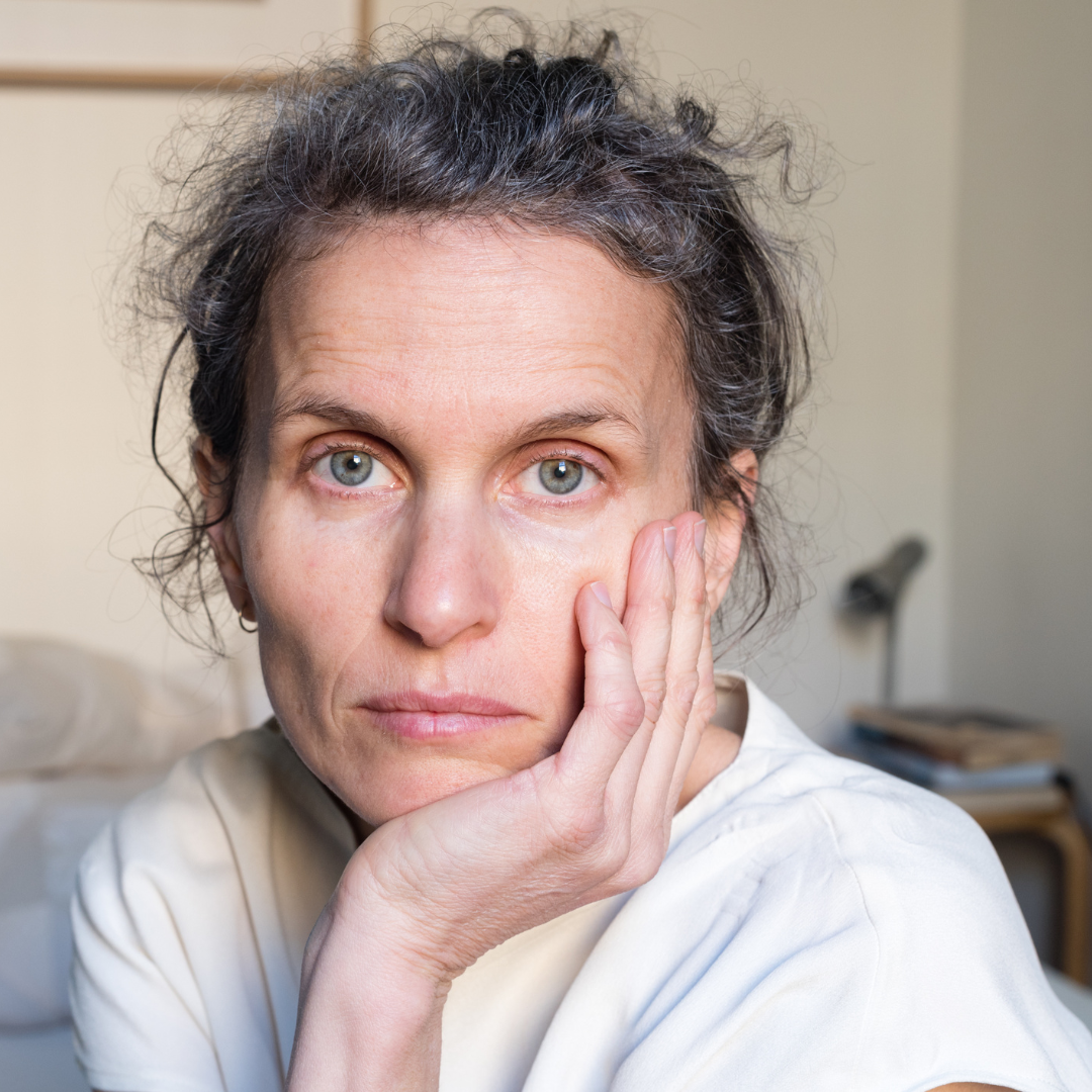 A woman with light skin and curly hair rests her face on her hand, looking thoughtfully at the camera. She wears a white top and sits in a softly lit room with a bed and books in the background.