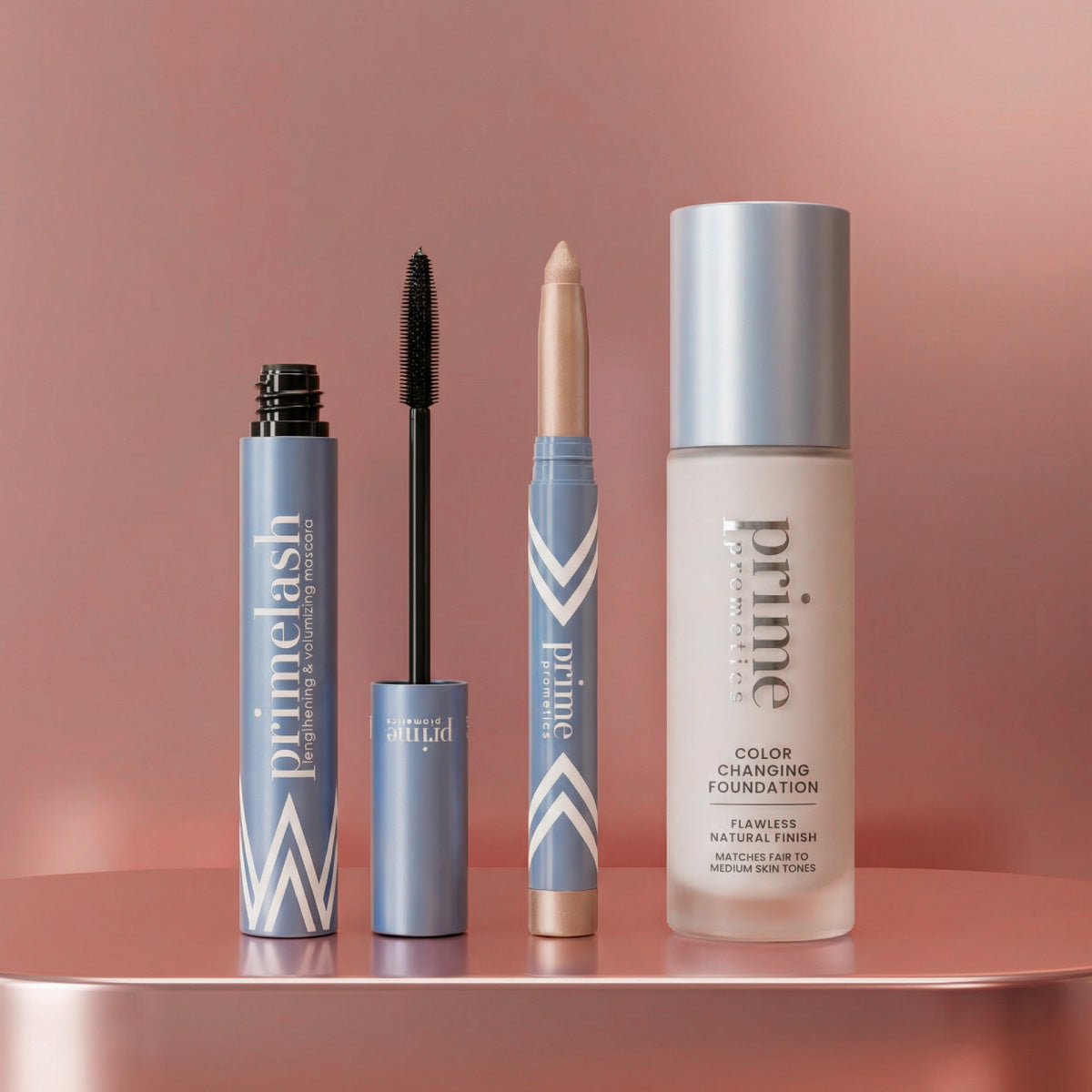 Three PrimePrometics™ Hollywood Glow Set products—a mascara, an eye crayon, and a color-changing foundation—stand upright on a glossy pink surface against a metallic pink background.