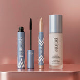 Three PrimePrometics™ Hollywood Glow Set products—a mascara, an eye crayon, and a color-changing foundation—stand upright on a glossy pink surface against a metallic pink background.
