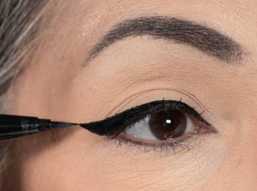 A close-up of a person’s eye as they apply black liquid eyeliner along the upper lid, creating a winged tip. The eyebrow above is dark and well-groomed.