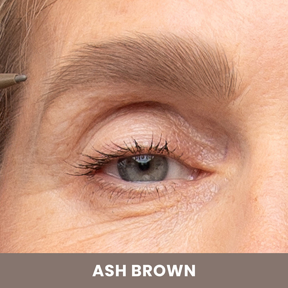 Close-up of a persons eye and eyebrow as a makeup pencil is applied to the eyebrow. The eyebrow color is labeled Ash Brown in text at the bottom of the image.