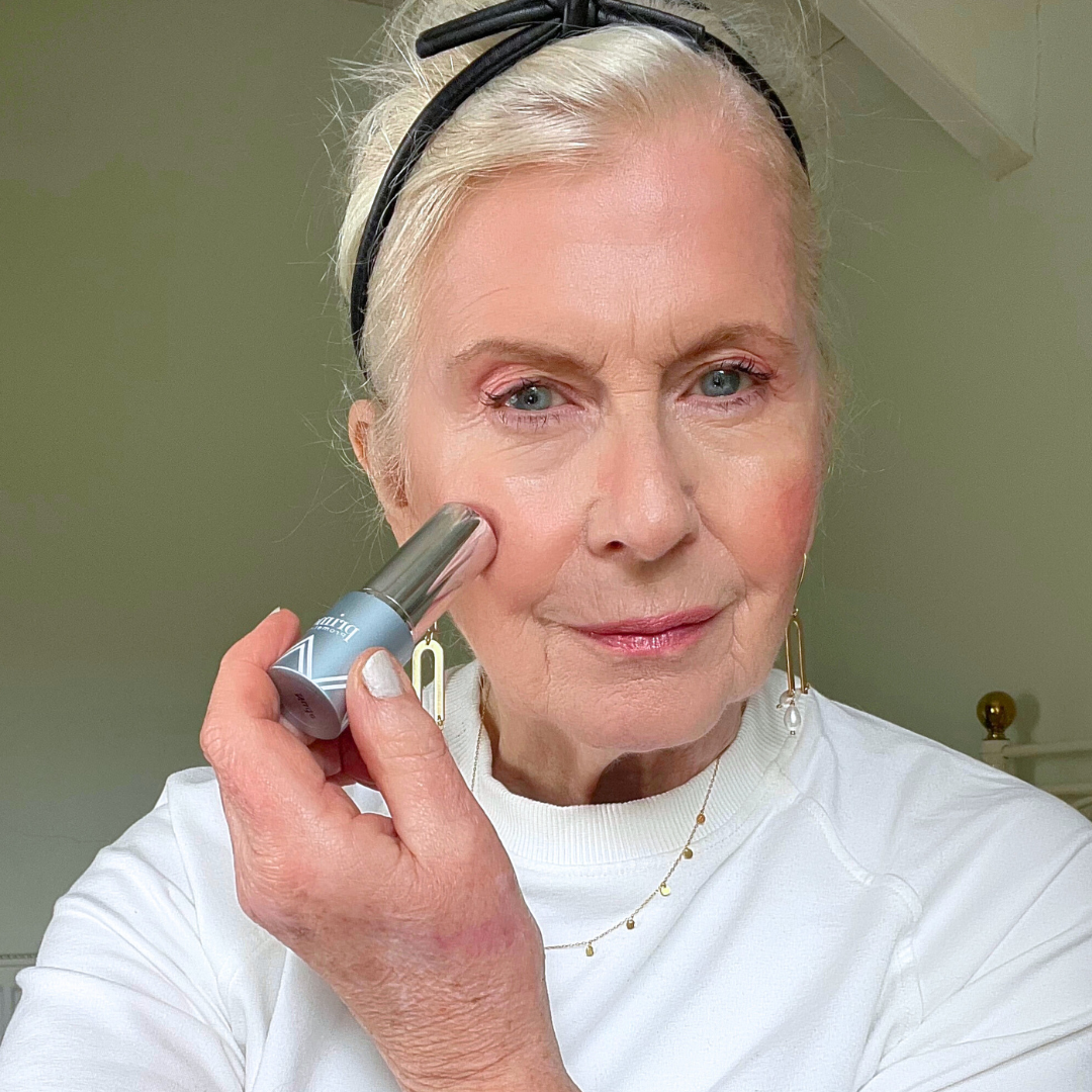An older woman with light skin and blonde hair, wearing a white top and black headband, applies makeup to her cheek using a stick applicator. She is looking at the camera in a softly lit room.