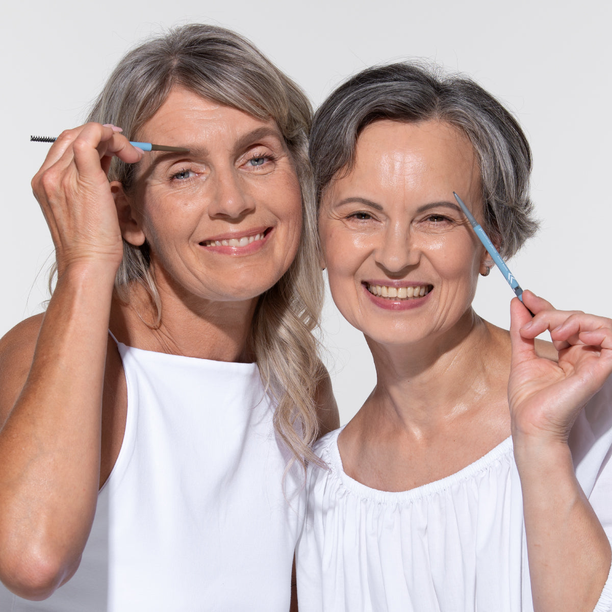 Two smiling older women with gray hair wearing white tops, posing together while shaping their eyebrows with blue eyebrow brushes against a plain light background.