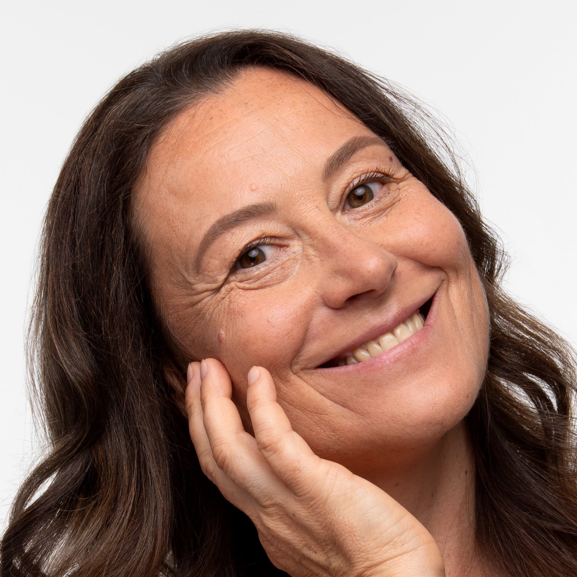 A middle-aged woman with long brown hair smiles warmly, resting her cheek on her hand against a plain white background.