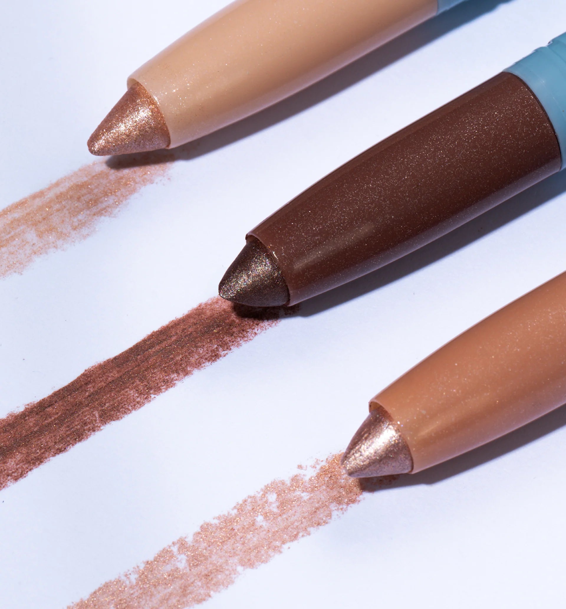 Three makeup pencils in nude, brown, and champagne shades are lined up horizontally, each drawing a matching color swatch on white paper. The pencils have metallic shimmery tips.