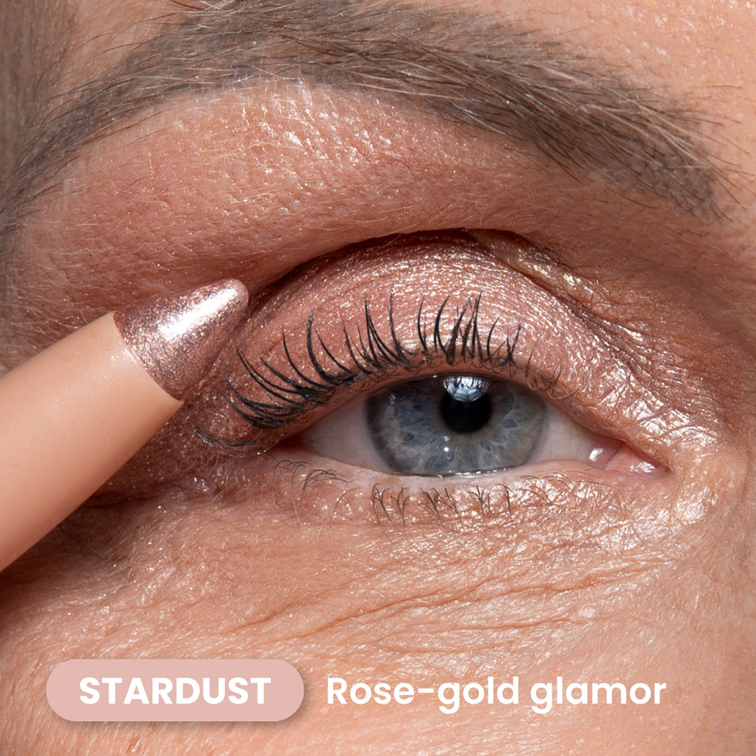 A close-up of a blue eye with shimmery rose-gold eyeshadow applied to the eyelid. A matching rose-gold makeup pencil touches the eyelid. Text: STARDUST Rose-gold glamor.
