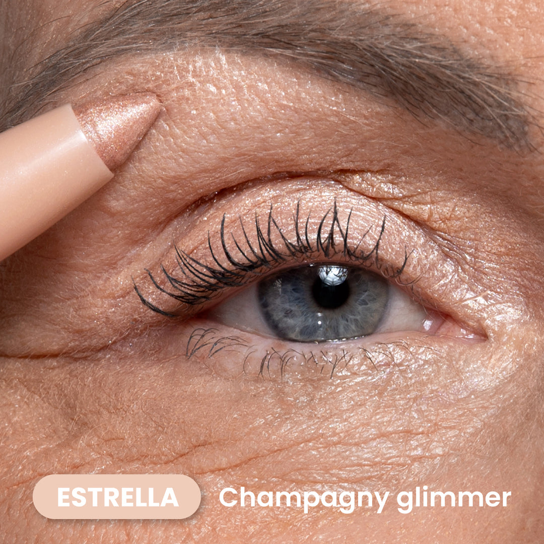 Close-up of a persons blue eye with shimmering champagne-colored eyeshadow and eyeliner applied. A pencil eyeshadow in a matching shade is held near the eyebrow. Text reads ESTRELLA Champagny glimmer.