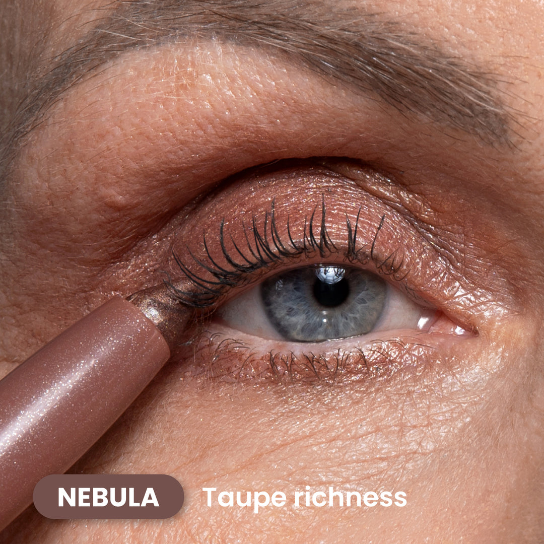 A close-up of a blue eye with taupe eyeshadow applied. A taupe eye pencil is being used on the upper eyelid. Text on the image reads NEBULA Taupe richness.