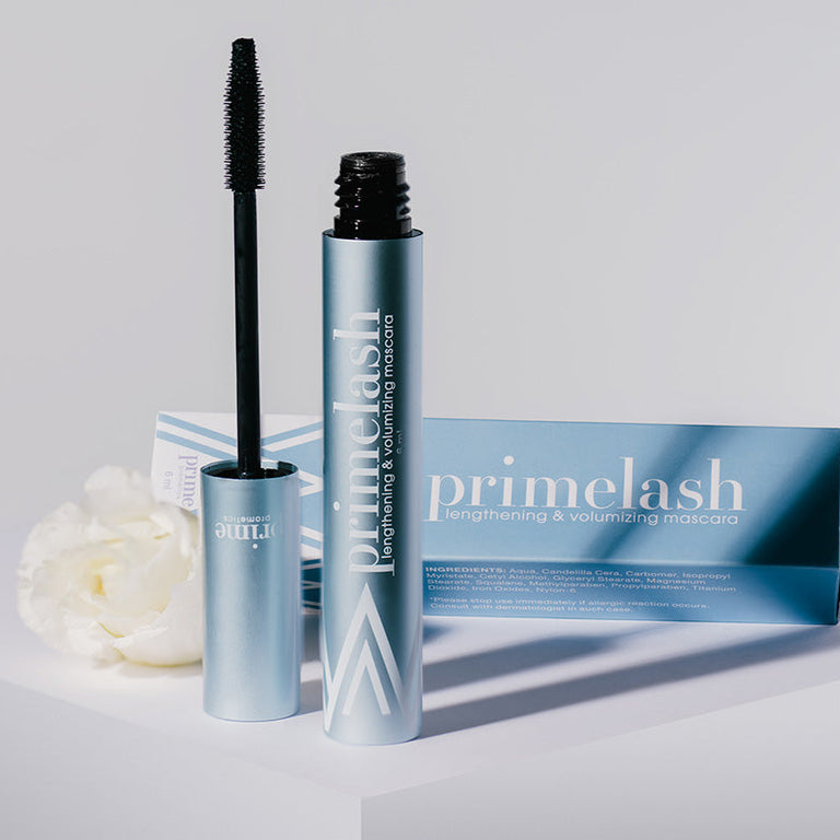 A blue tube of Primelash lengthening & volumizing mascara with the wand applicator is standing upright next to its blue box packaging, placed on a white surface with a white flower nearby.