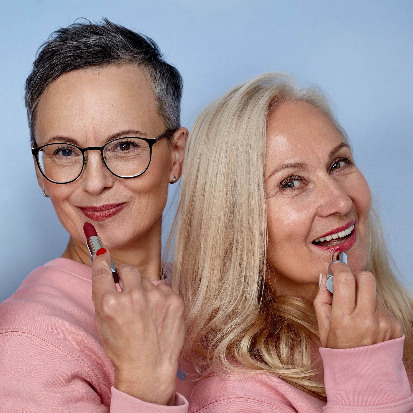 Two smiling older women with short gray hair and long blonde hair, both wearing pink tops, stand back to back while applying red lipstick, against a light blue background.