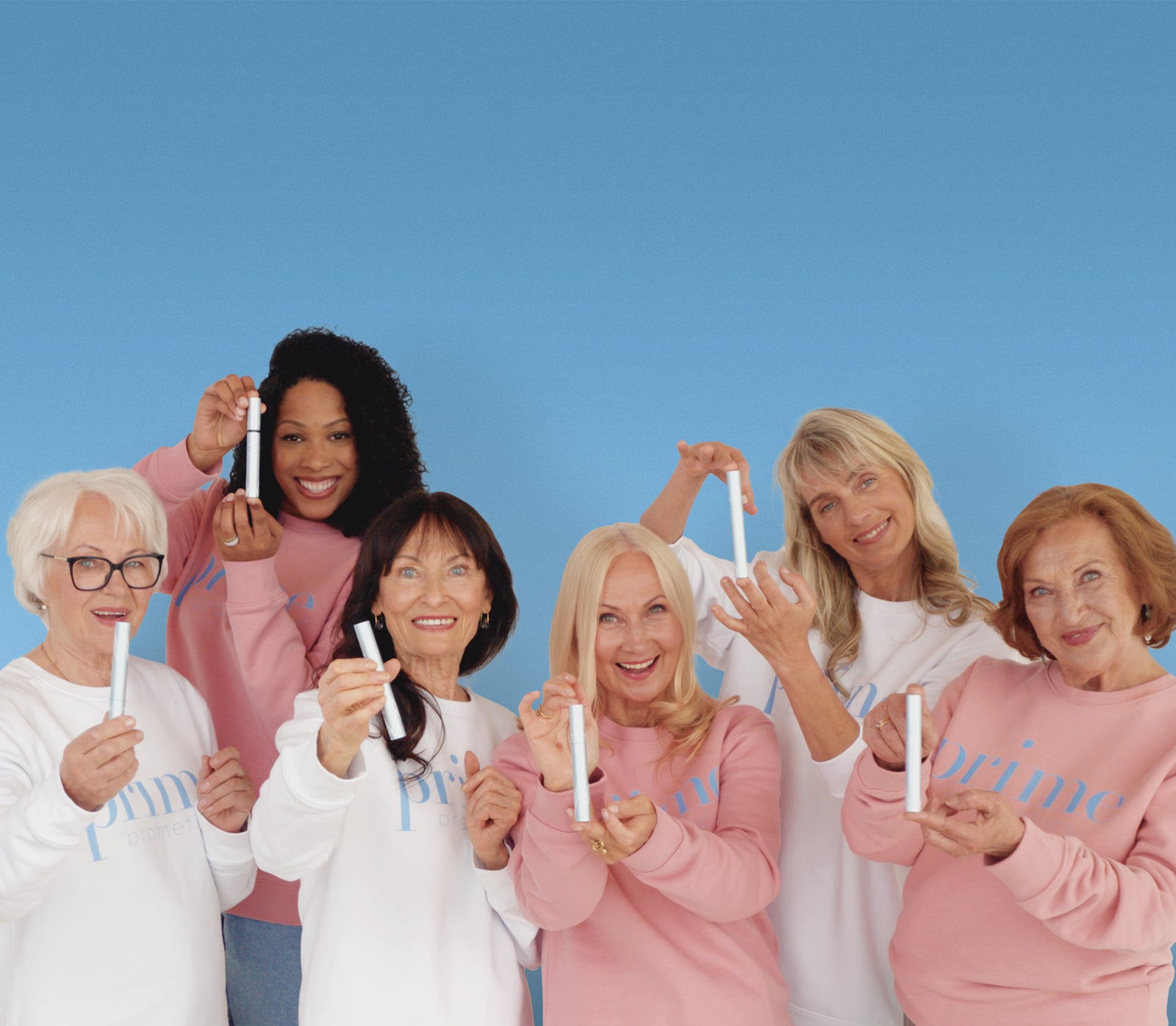 Six smiling older women wearing pink and white prime sweatshirts, standing together against a blue background, each holding up a white beauty product or makeup item toward the camera.