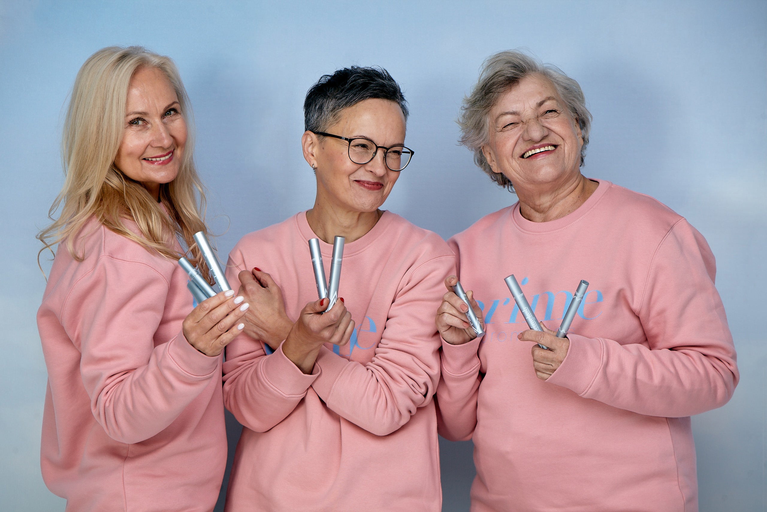 Three smiling older women wearing matching light pink sweatshirts hold several silver cosmetic tubes against a light blue background. They stand close together, looking cheerful and confident.