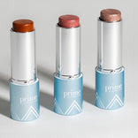 Three silver and blue tubes of Prime Prometics makeup sticks are standing upright. Each stick has a different shade: brown, pink, and shimmery bronze. The background is a plain, light surface.