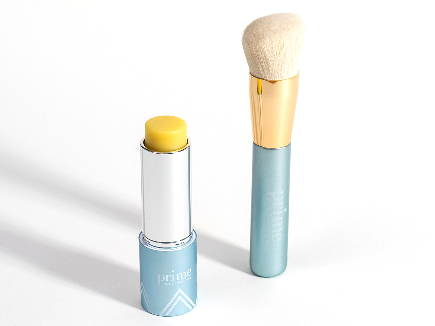 A blue and silver tube of yellow lip balm labeled Prime stands open next to a matching blue and gold makeup brush with soft, light bristles, both on a white background.