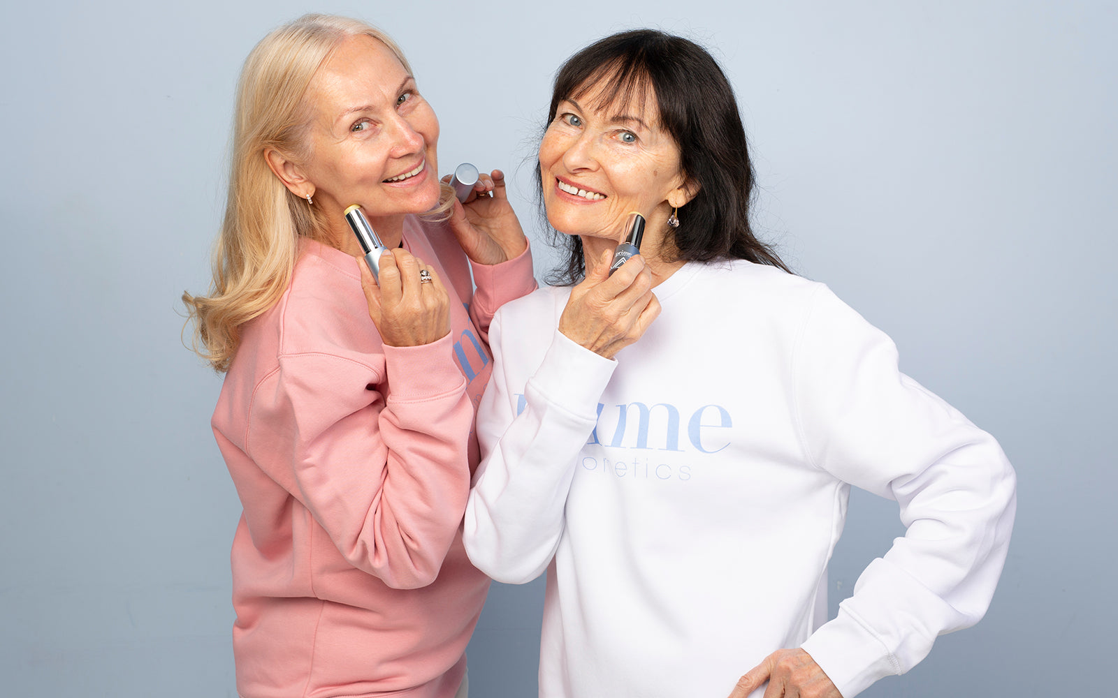 Two smiling older women in pastel sweaters apply makeup sticks to their faces while standing side by side against a light blue background.