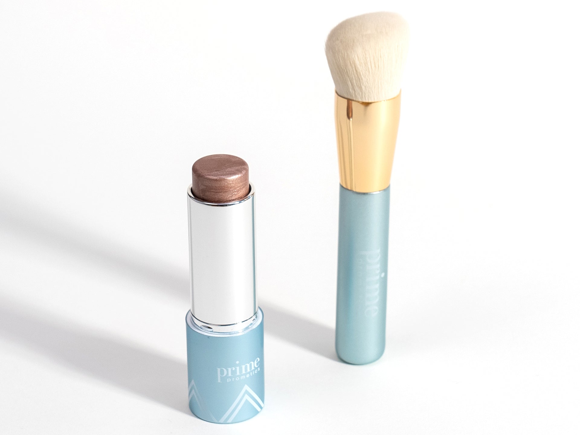 A makeup stick with a metallic rose shade in a blue tube labeled prime sits next to a makeup brush with a gold and blue handle and cream bristles on a white background.