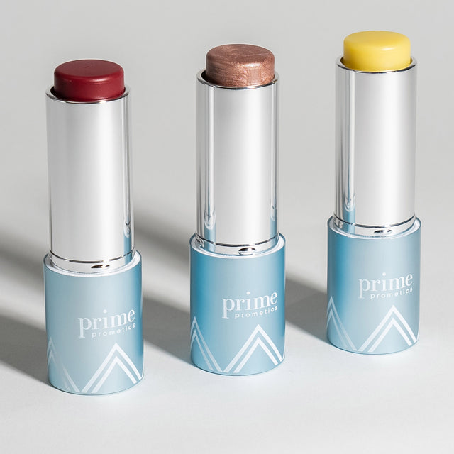Three PrimePrometics™ PrimeWand Trinity sticks stand upright, each in a silver case with a blue base and white geometric design, showing multitasking shades: yellow, deep red, and shimmery beige.