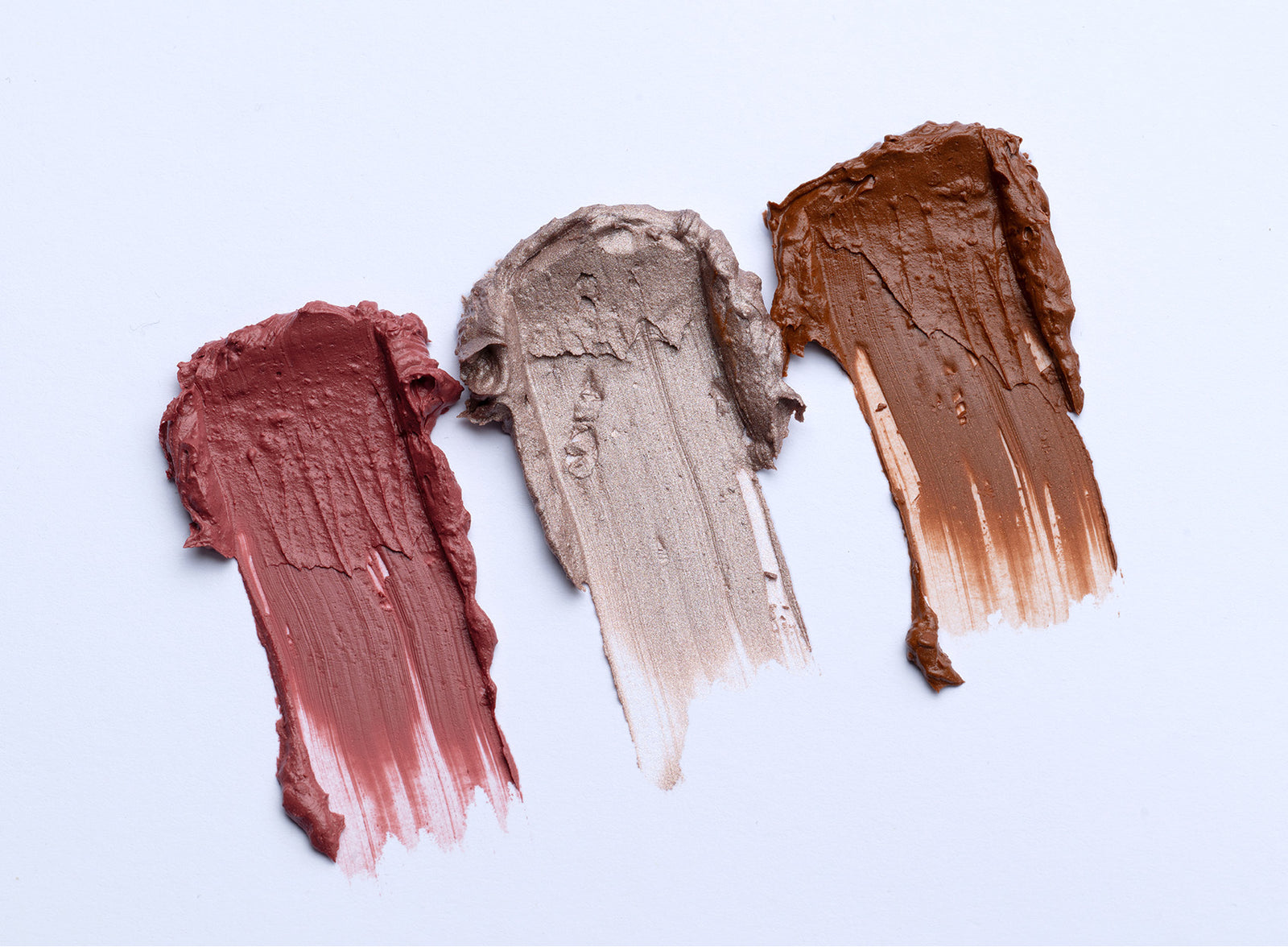Three swatches of creamy makeup in mauve, metallic taupe, and brown shades are smeared side by side on a white background.
