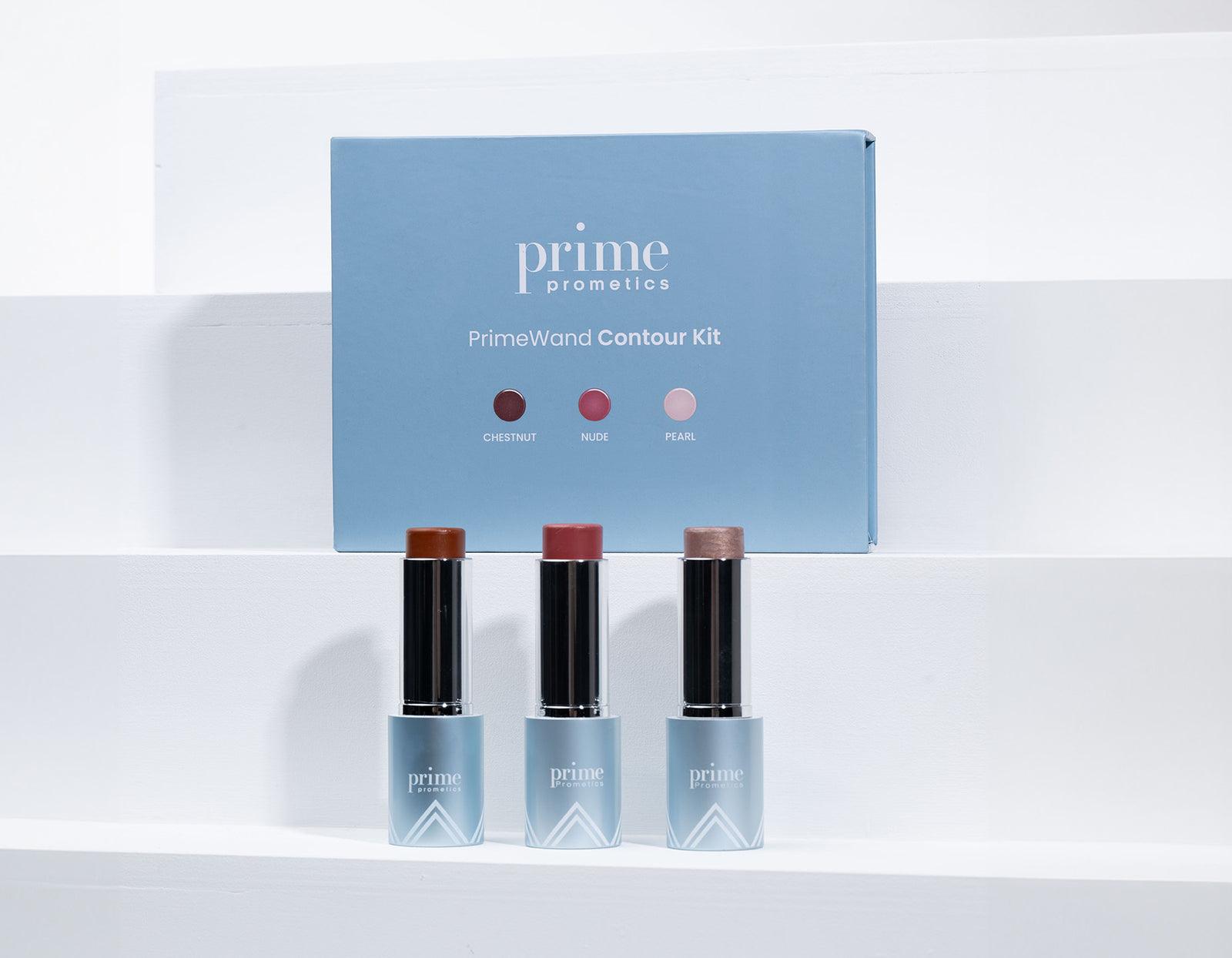 Three Prime Prometics makeup sticks—chestnut, nude, and pearl—stand in front of a blue PrimeWand Contour Kit box, displayed on white steps.