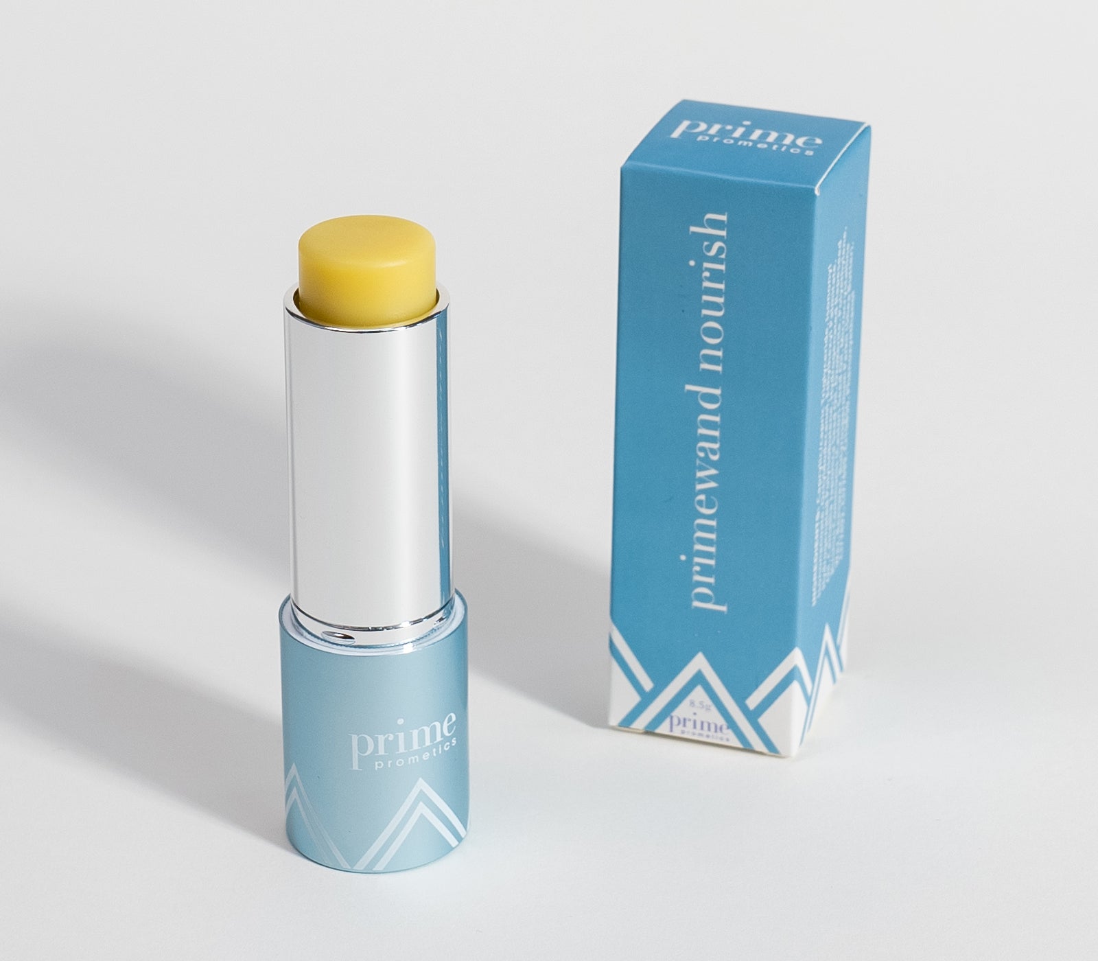 A blue tube of Prime Prometics PrimeWand Nourish balm with its cap off, showing a yellow balm stick, is placed next to its matching blue and white product box on a light background.
