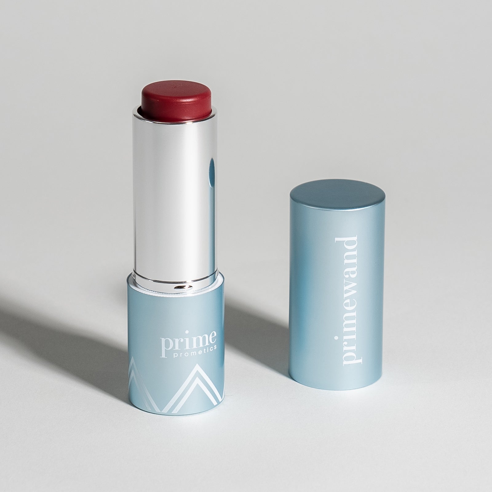 A silver and blue lipstick tube with a deep red lipstick exposed, standing upright next to its matching blue cap labeled prime wand on a light gray surface.