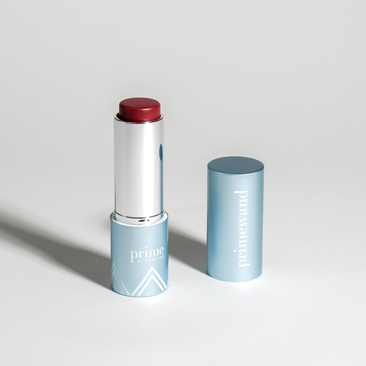 A blue tube of lipstick with the cap off, showing a deep red shade. The tube is labeled prime and primewand, and both the tube and cap are placed on a light gray surface.