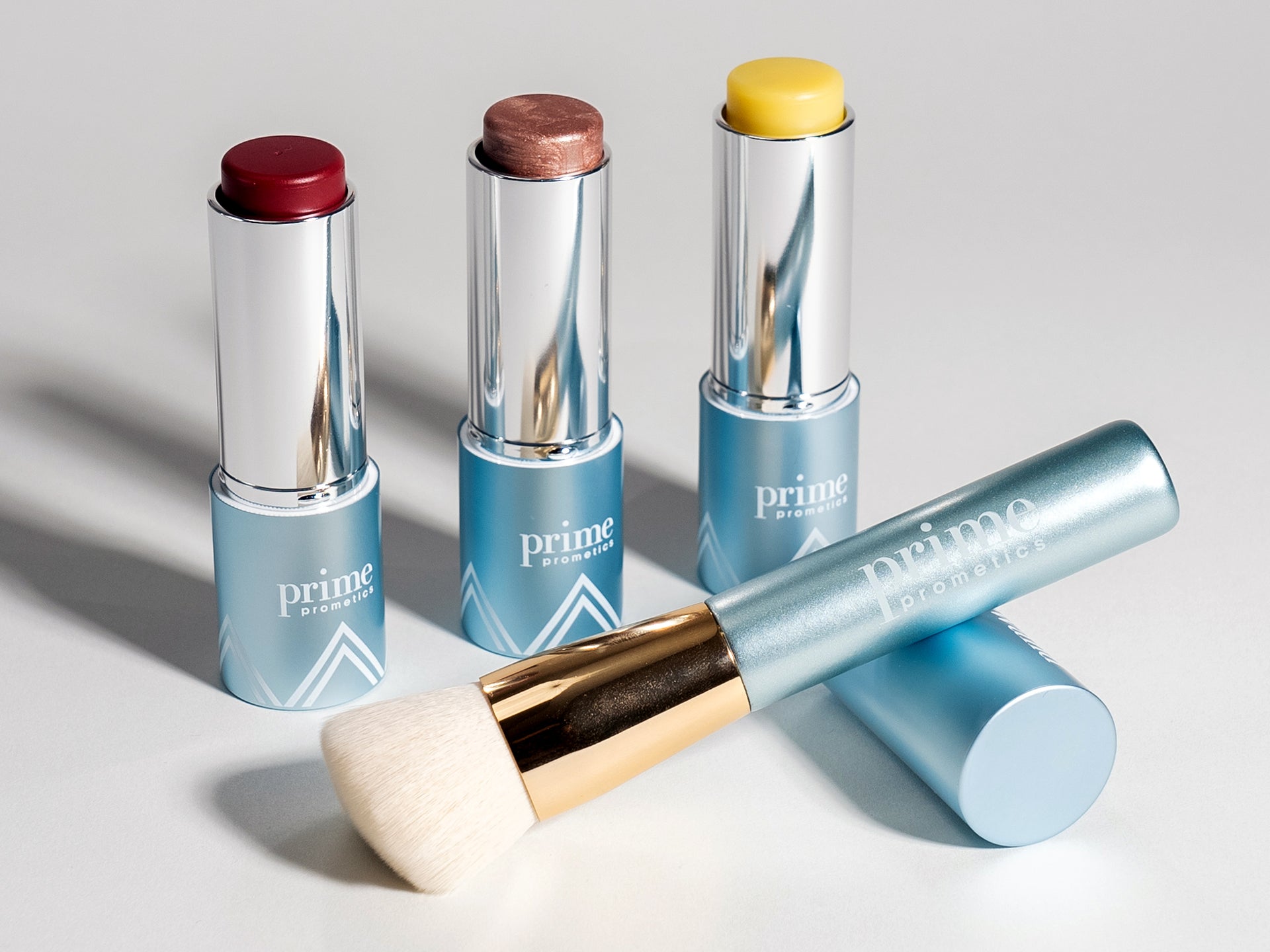 Three open makeup sticks—one red, one shimmery bronze, and one yellow—stand upright next to a blue tube labeled prime prometics with a matching makeup brush attached. All items have metallic accents.