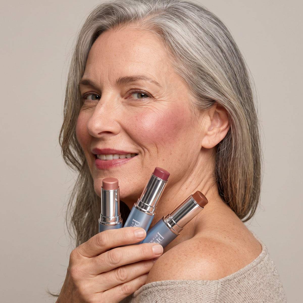 A smiling older woman with gray hair holds three PrimePrometics™ PrimeWands 5-in-1 Makeup Wands in different shades near her face. Wearing a beige off-the-shoulder sweater and a natural makeup look, she demonstrates the versatility and buildable color of this multi-use makeup wand, all set against a neutral background.