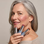 A smiling older woman with gray hair holds three PrimePrometics™ PrimeWands 5-in-1 Makeup Wands in different shades near her face. Wearing a beige off-the-shoulder sweater and a natural makeup look, she demonstrates the versatility and buildable color of this multi-use makeup wand, all set against a neutral background.