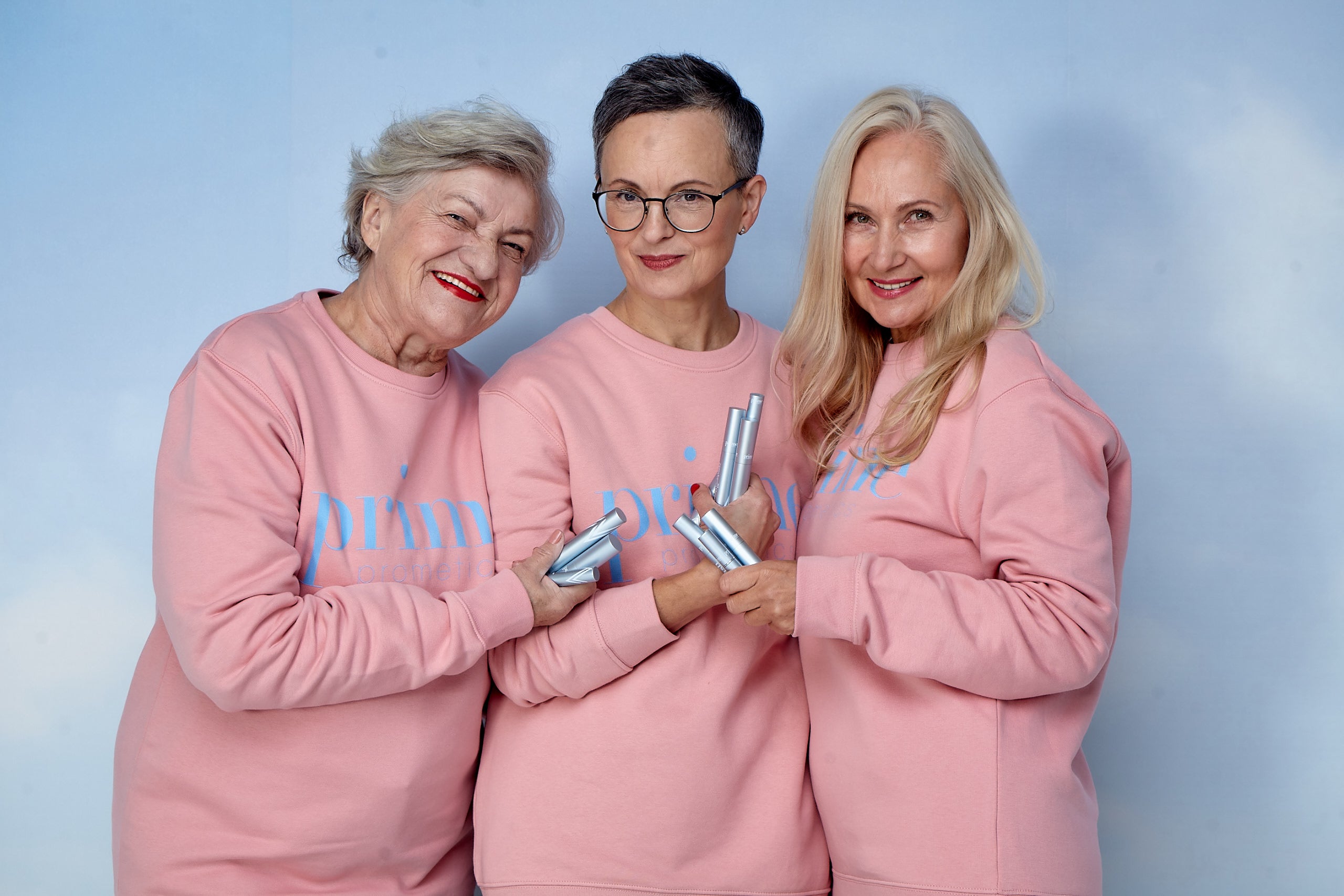 Three older women wearing matching pink sweatshirts stand together smiling, holding silver cosmetic bottles against a light blue background.