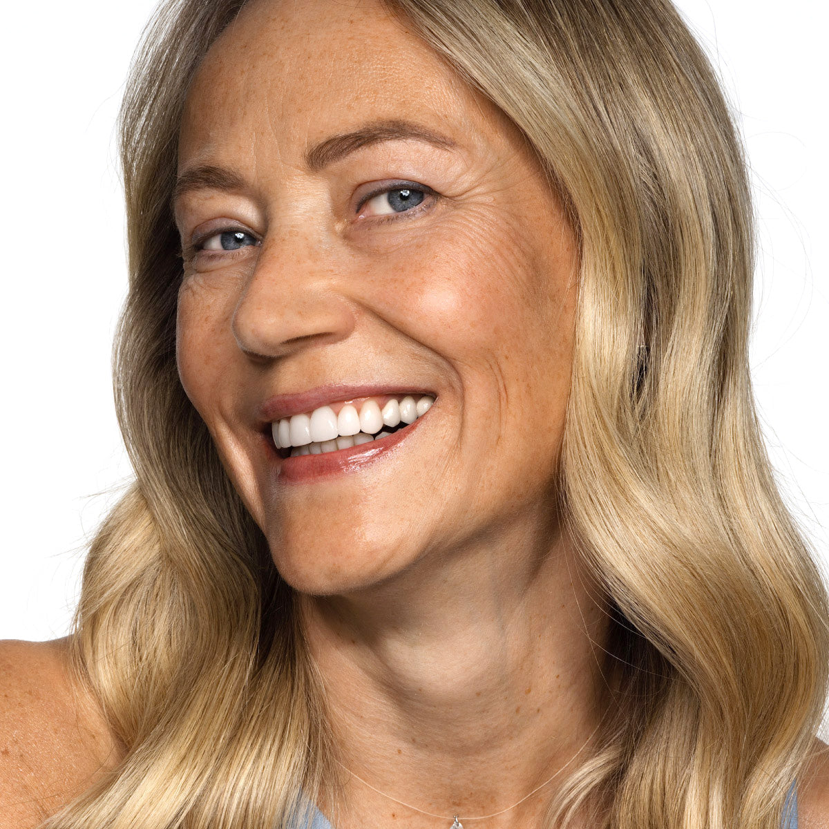 A woman with long, wavy blonde hair smiles warmly, showing her teeth. She has blue eyes, light skin with freckles, and is photographed against a plain white background.