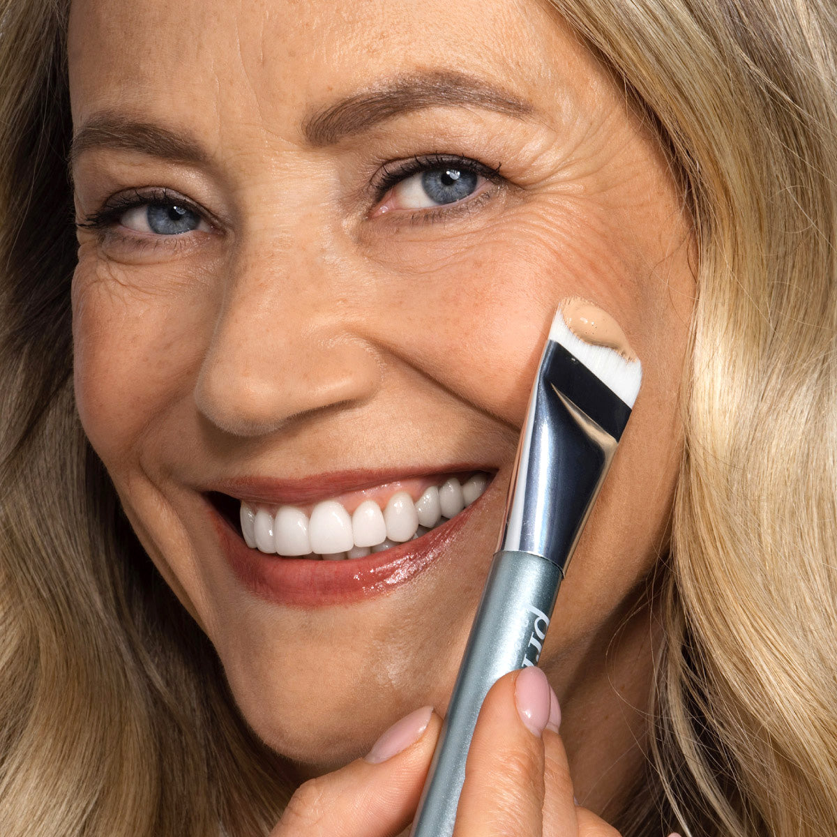 A smiling woman with blonde hair applies cream foundation to her cheek using a makeup brush. She has blue eyes, clear skin, and is looking towards the camera.
