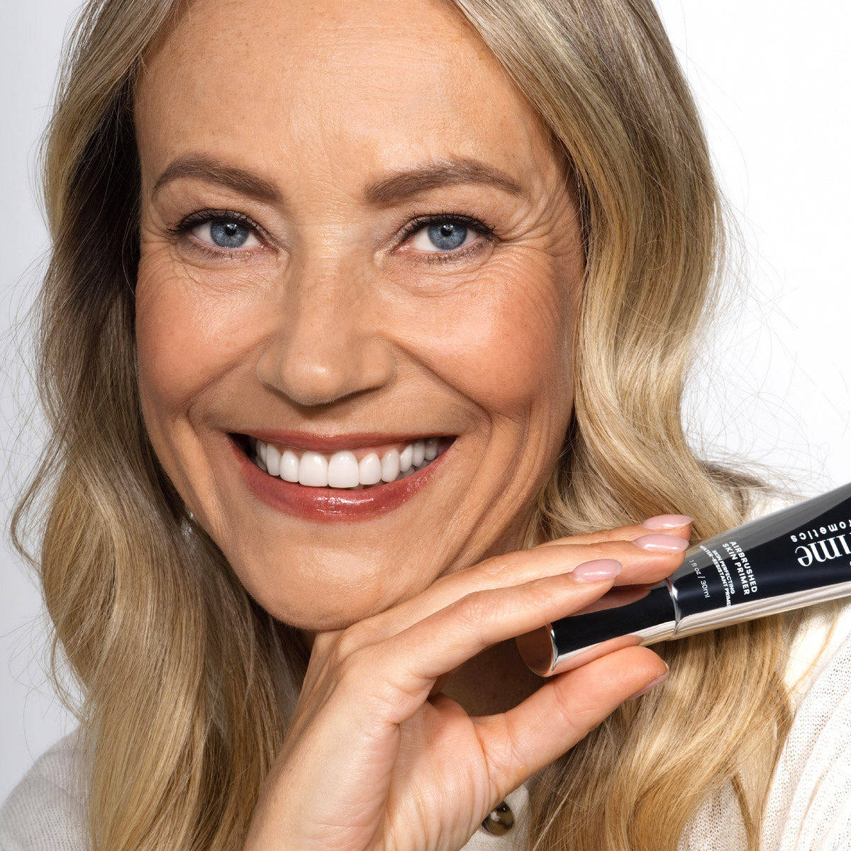 A smiling woman with long blonde hair holds a black and silver cosmetic product near her face, showing clear skin and white teeth.